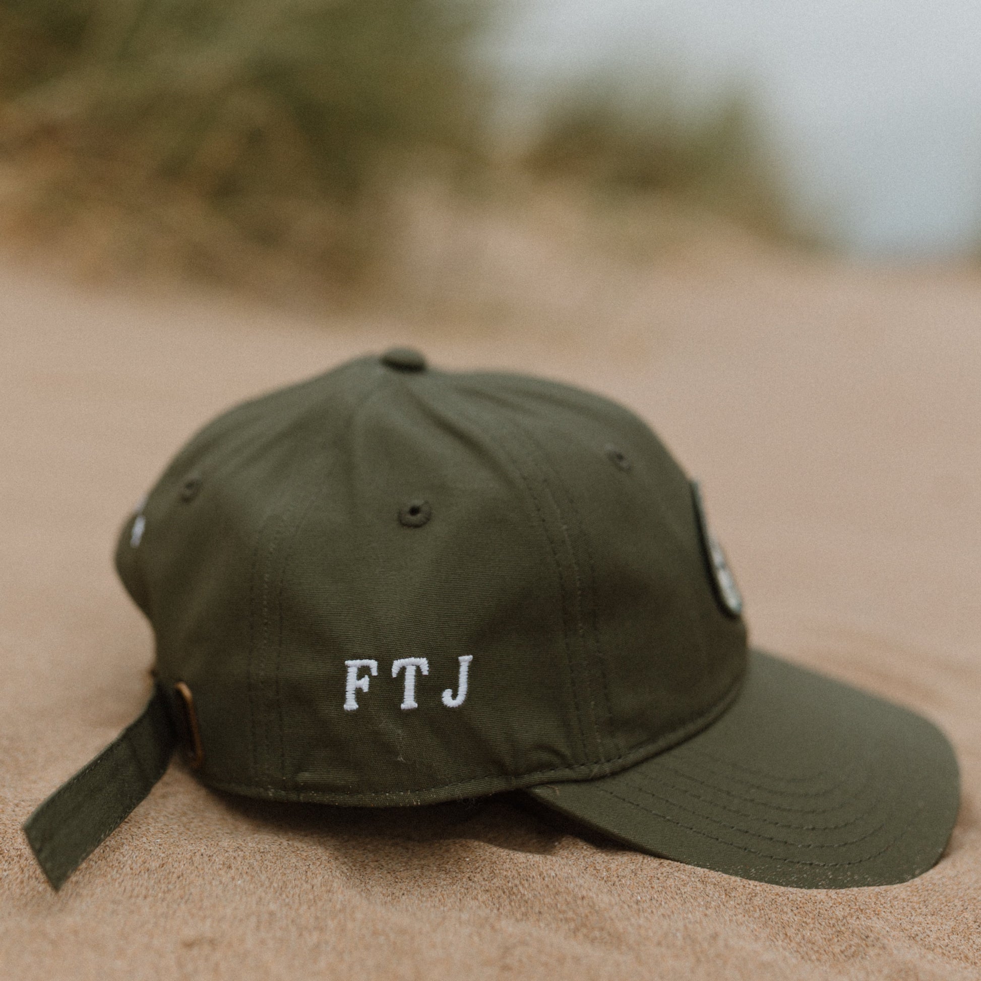 A khaki green baseball cap with white embroidered letters 'FTJ' on the front, resting on a sandy beach with blurred dune grass in the background.