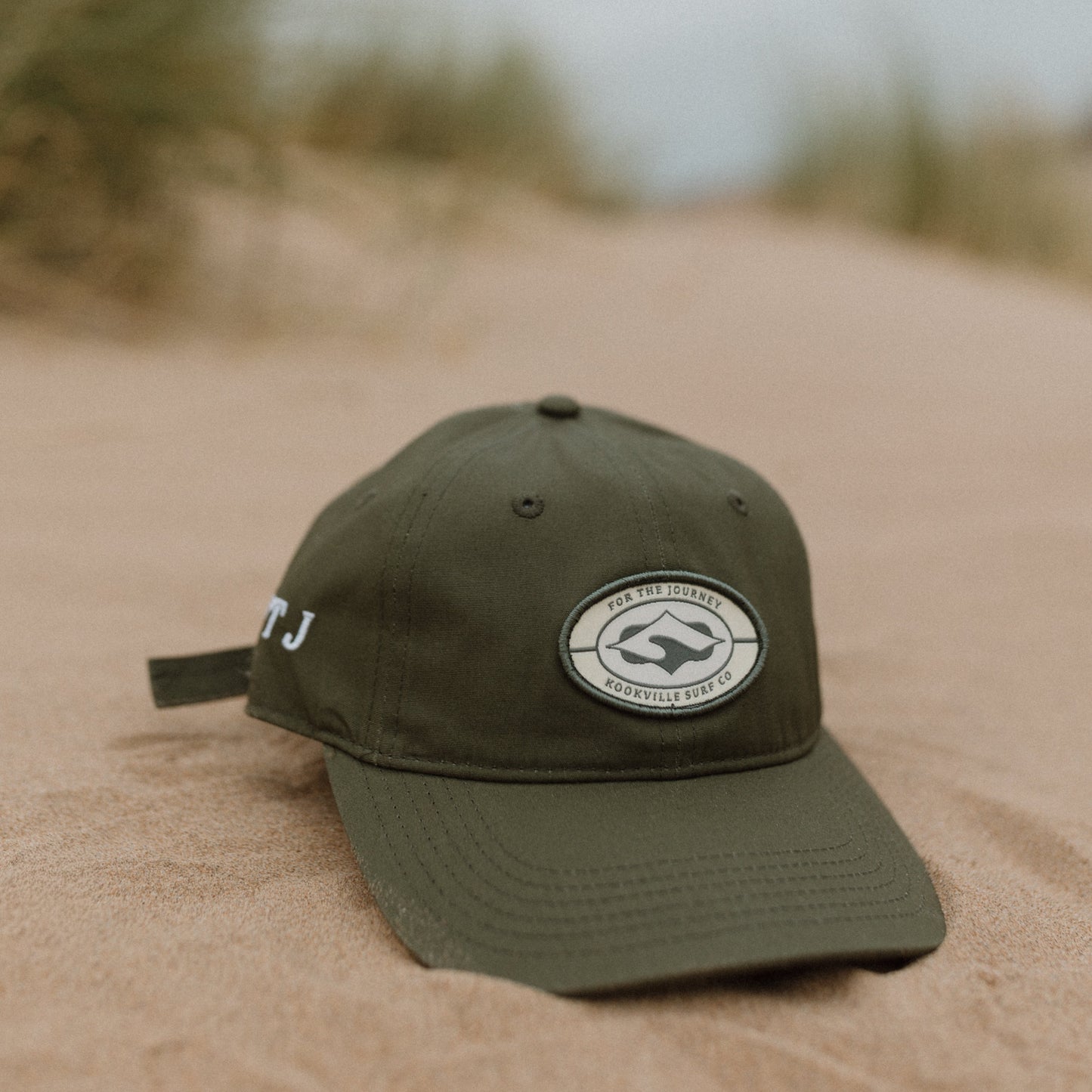 Khaki green baseball cap resting on sand in an outdoor dune setting, featuring a Kookville Surf Co circular white embroidered logo patch on the front and white lettering on the side.